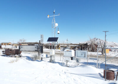 Snow-covered weather monitoring station with solar panels, sensors, and satellite dishes, set in a rural area with wooden structures and mountains in the background.