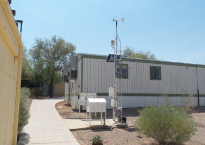 A small weather station with instruments and a tall metal tower stands outside a modular building in a dry, desert-like environment with shrubs and a clear sky.