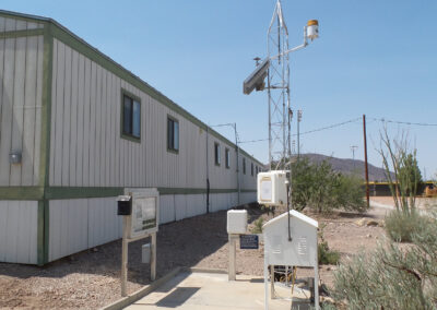 Portable weather station equipment installed on concrete next to a long, light-colored modular building in a dry, desert environment.