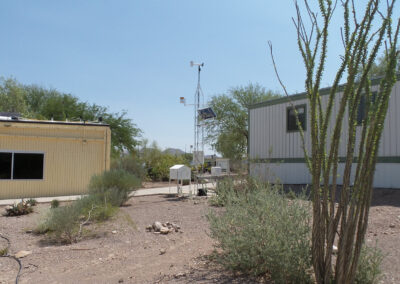 Two single-story buildings with light-colored exteriors surround a weather station and sparse desert vegetation under a clear sky.