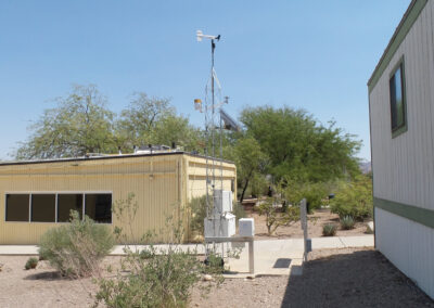 A small weather station with sensors and anemometer stands beside a yellow building and a mobile unit amid desert vegetation under a clear sky.