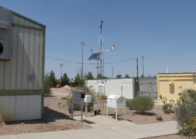 Weather monitoring equipment and sensors are installed outdoors near modular buildings, with a solar panel and antenna mounted on a tall pole under a clear sky.