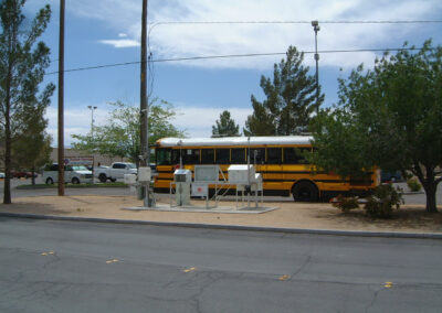 A yellow school bus is parked in a lot next to utility boxes, with trees and cars in the background under a partly cloudy sky.