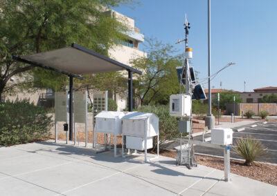 Outdoor weather station with various meteorological instruments, sensors, and equipment, located near a parking lot and surrounded by small desert plants.