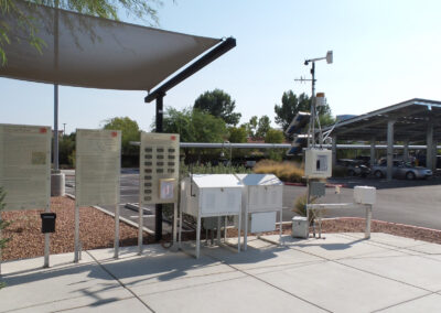 Outdoor weather monitoring station with multiple information boards and meteorological instruments, located near a parking lot with solar panel covers and surrounding vegetation.