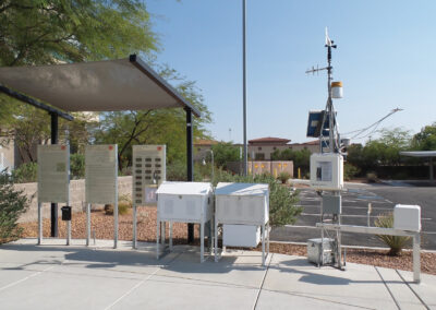A weather station with various meteorological instruments, information boards, and solar panels is set up on a paved area surrounded by desert landscaping.