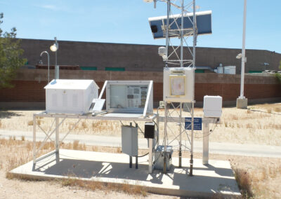 Weather monitoring station with various instruments, solar panels, and sensors mounted on a metal structure in a dry, outdoor setting near a building.