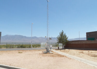 A weather station with sensors and anemometer is installed on bare ground near a sidewalk, with mountains and a building visible in the background under a clear sky.