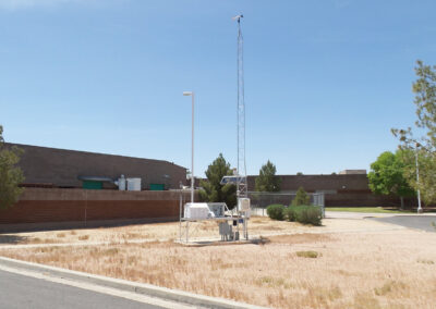 A weather monitoring station with sensors and an antenna stands on dry grass near a brick wall and trees under a clear sky.