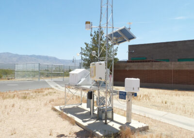 A weather monitoring station with sensors and solar panels is set up on dry ground near a fenced area and brick building under a clear sky.