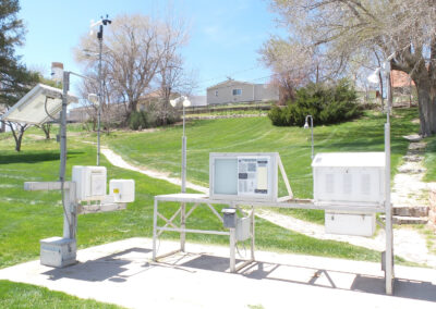 Outdoor weather monitoring station with multiple instruments and sensors mounted on metal stands, set on a concrete pad in a grassy area with trees and buildings in the background.