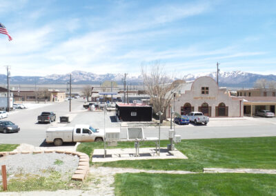 Small town street with vehicles parked, a restaurant building, an American flag, and mountains in the background under a clear sky.