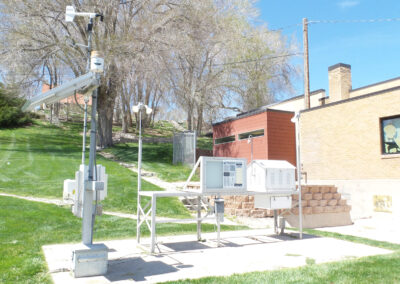 Outdoor weather monitoring station with various sensors and equipment installed on a metal frame, situated on grass near a building and trees.