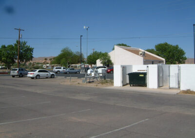 A parking lot with several parked cars, a white building, a green dumpster in an enclosure, and trees under a clear blue sky.