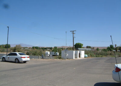 Two parked cars in an empty asphalt lot near a small white utility building with storage tanks, power lines, and distant trees under a clear blue sky.