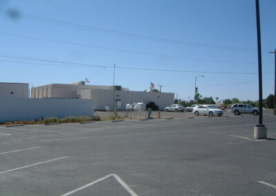 A mostly empty parking lot with a few cars parked near a low, white building. The sky is clear and blue, and several American flags are visible on the building.