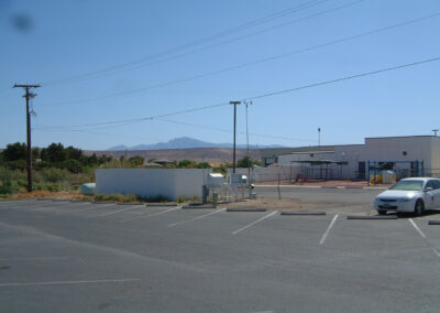 Empty parking lot with a few parked cars, a fenced playground, utility structures, and a white building; mountains and clear sky in the background.