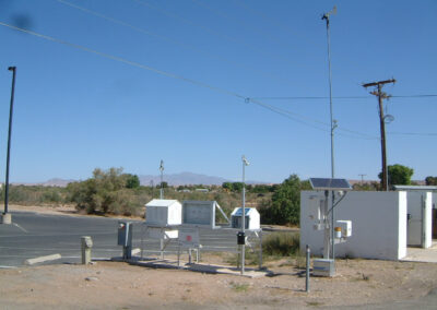 A weather monitoring station with instruments, a solar panel, and a white enclosure is set up beside a parking area in a dry, desert landscape under a clear blue sky.