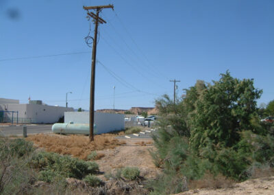 A telephone pole and power lines run alongside a road near a white building, with dry vegetation and a propane tank visible in the foreground.