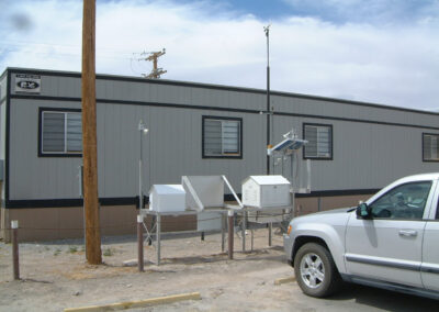A weather monitoring station with various instruments is set up beside a gray building and a white vehicle in a gravel lot.