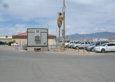Parking lot with several cars, a small building, a palm tree, and mountains visible in the background under a cloudy sky. A person walks on the left side.