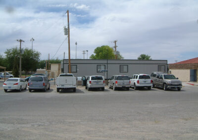 Six vehicles are parked in a row in front of a gray, rectangular, single-story building on a cloudy day. The lot is paved, and utility poles are visible in the background.