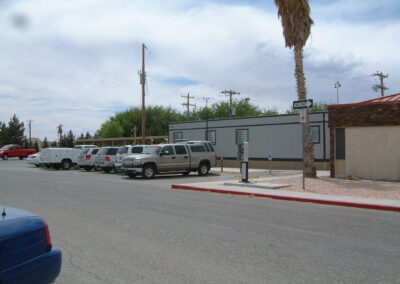 A row of parked cars beside a gray modular building on a paved street, with a payphone, palm tree, and utility poles visible under a partly cloudy sky.
