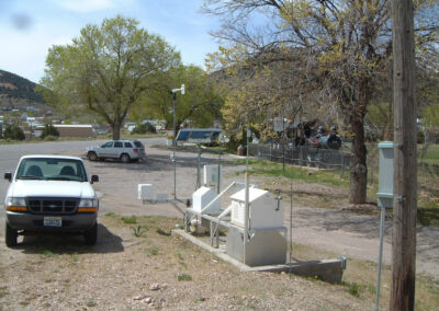 A white pickup truck is parked near a weather monitoring station with various sensors and equipment. Trees, a fence, and parked vehicles are visible in the background.