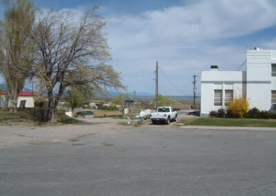 A white truck is parked near a white building on a mostly empty street, with a few trees and utility poles under a partly cloudy sky.