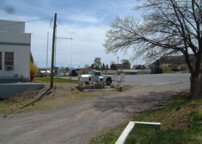 A white pickup truck is parked on a gravel road near utility equipment, with a building and houses in the background under a partly cloudy sky.