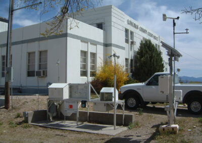 A weather monitoring station with various instruments is set up outside a white government building, with a white pickup truck parked nearby.