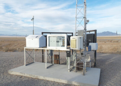 An outdoor air quality monitoring station with various instruments and sensors is set up on a concrete pad in a desert landscape.