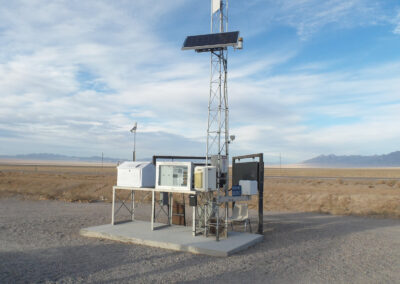 A weather monitoring station with solar panels stands on a concrete platform in a desert landscape with mountains visible in the distance.
