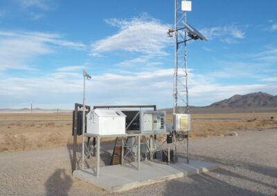 A ground-based weather station with various sensors and instruments stands on a concrete slab in a dry, open landscape with mountains and a road in the background.