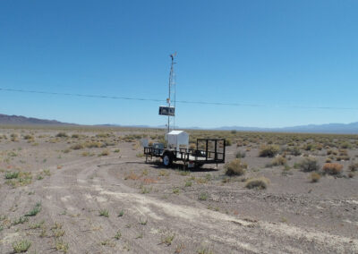 A mobile weather station on a trailer is positioned in a dry, open desert landscape under a clear blue sky.