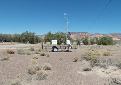 A weather monitoring station mounted on a trailer sits in an arid landscape with sparse vegetation and distant hills under a clear blue sky.