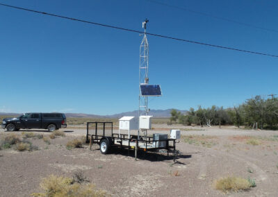 A mobile weather station with solar panels stands in a dry, open landscape near a parked black SUV under a clear blue sky.