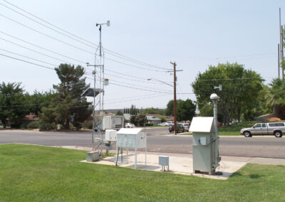 Outdoor weather monitoring station with various meteorological instruments and equipment beside a street, grass lawn in foreground, cars and trees in background.