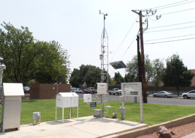 Outdoor weather monitoring station with various instruments, sensors, and a solar panel on a grassy area near a parking lot and trees.