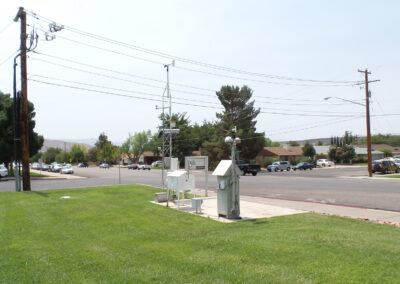 A small weather monitoring station with sensors and equipment is positioned on a concrete pad next to a grassy area by a street in a suburban neighborhood.