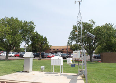 Outdoor weather monitoring station with metal equipment, solar panel, antenna, and sensors on a grassy area, parking lot and building visible in the background.