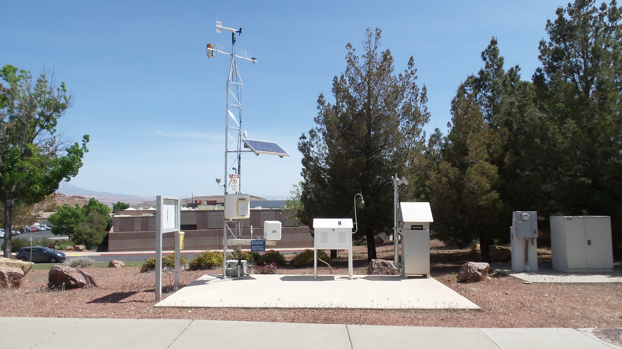 A weather station with various meteorological instruments, including a solar panel, sensors, and weather tower, is set up on a concrete pad surrounded by trees and buildings.