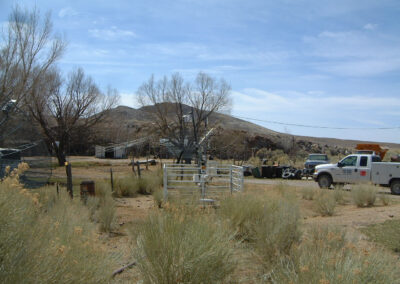 A rural, desert landscape with dry shrubs, leafless trees, fencing, utility truck, and small buildings in the background under a partly cloudy sky.