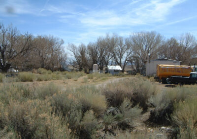 A rural landscape with dry shrubs, leafless trees, a few small buildings, and a yellow tanker trailer near an old vehicle under a blue sky.