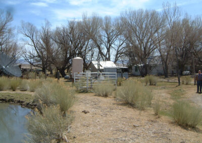 A rural yard with dry grass, shrubs, bare trees, a pond, metal gates, various structures, and one person standing at the right edge.