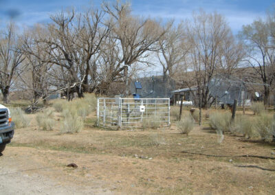 A fenced weather station with sensors and solar panel stands in a dry, grassy area with leafless trees and satellite dishes in the background.