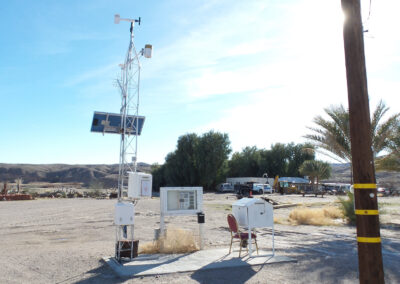 A weather monitoring station with solar panels and sensors is set up in a desert area near utility poles, trees, and distant vehicles under a clear sky.