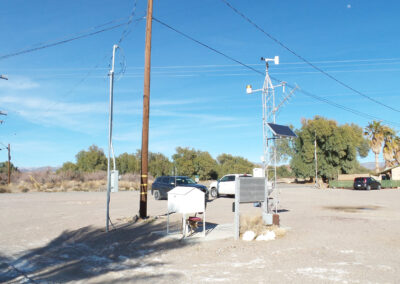 A weather monitoring station with sensors, solar panel, and instruments stands in a dry, open area with parked cars, power lines, and trees in the background.