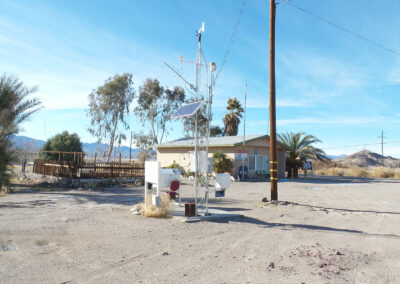 A weather monitoring station stands in a dry, open area near a small building, surrounded by scattered palm trees and power lines under a clear blue sky.