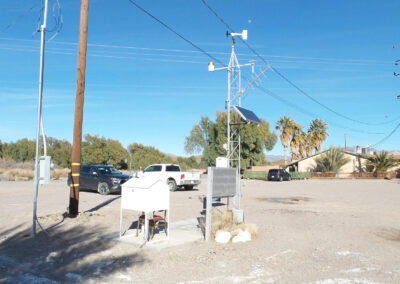 Weather monitoring equipment and electrical boxes are installed on a gravel lot with parked vehicles, utility poles, trees, and clear blue sky in the background.